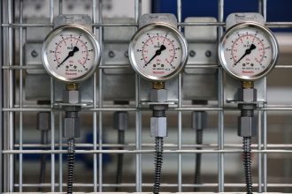 Close de manômetros industriais instalados em uma grade metálica dentro de uma fábrica.