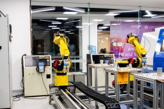 Braços robóticos operam em um ambiente industrial moderno, com equipamentos de controle ao fundo.