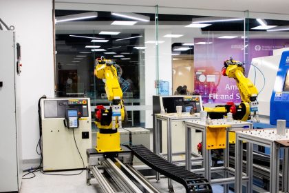 Braços robóticos operam em um ambiente industrial moderno, com equipamentos de controle ao fundo.