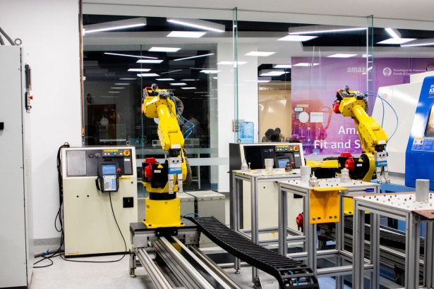 Braços robóticos operam em um ambiente industrial moderno, com equipamentos de controle ao fundo.