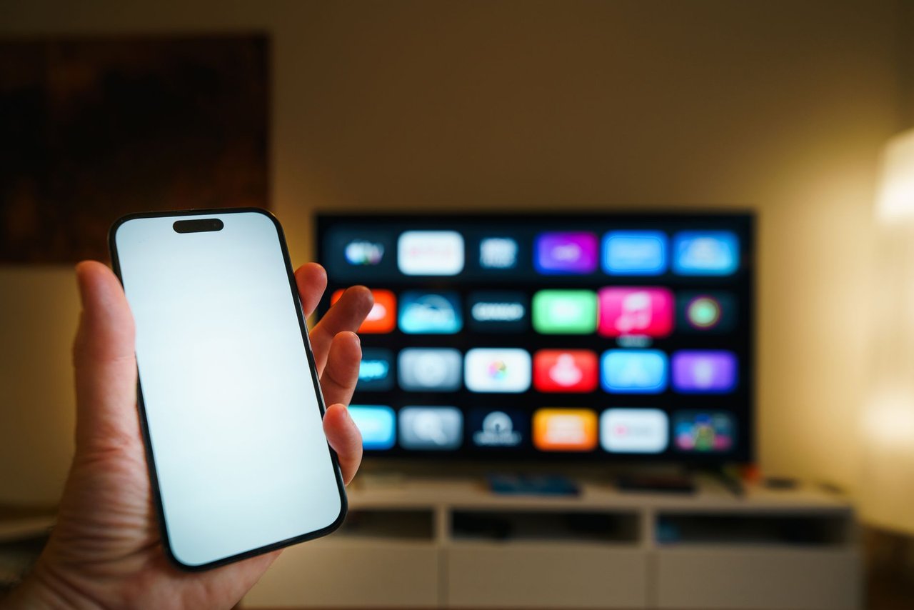 Smartphone em primeiro plano diante de uma televisão inteligente com aplicativos exibidos em uma sala de estar.