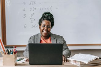 Professora sentada à mesa de uma sala de aula, trabalhando diante de um netbook.