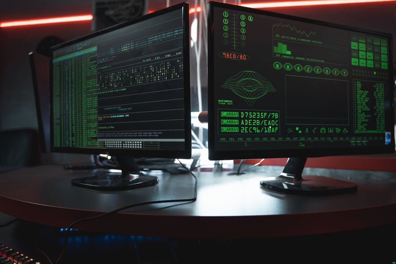 Dois monitores exibem interfaces de código em uma sala escura, em referência à segurança digital.